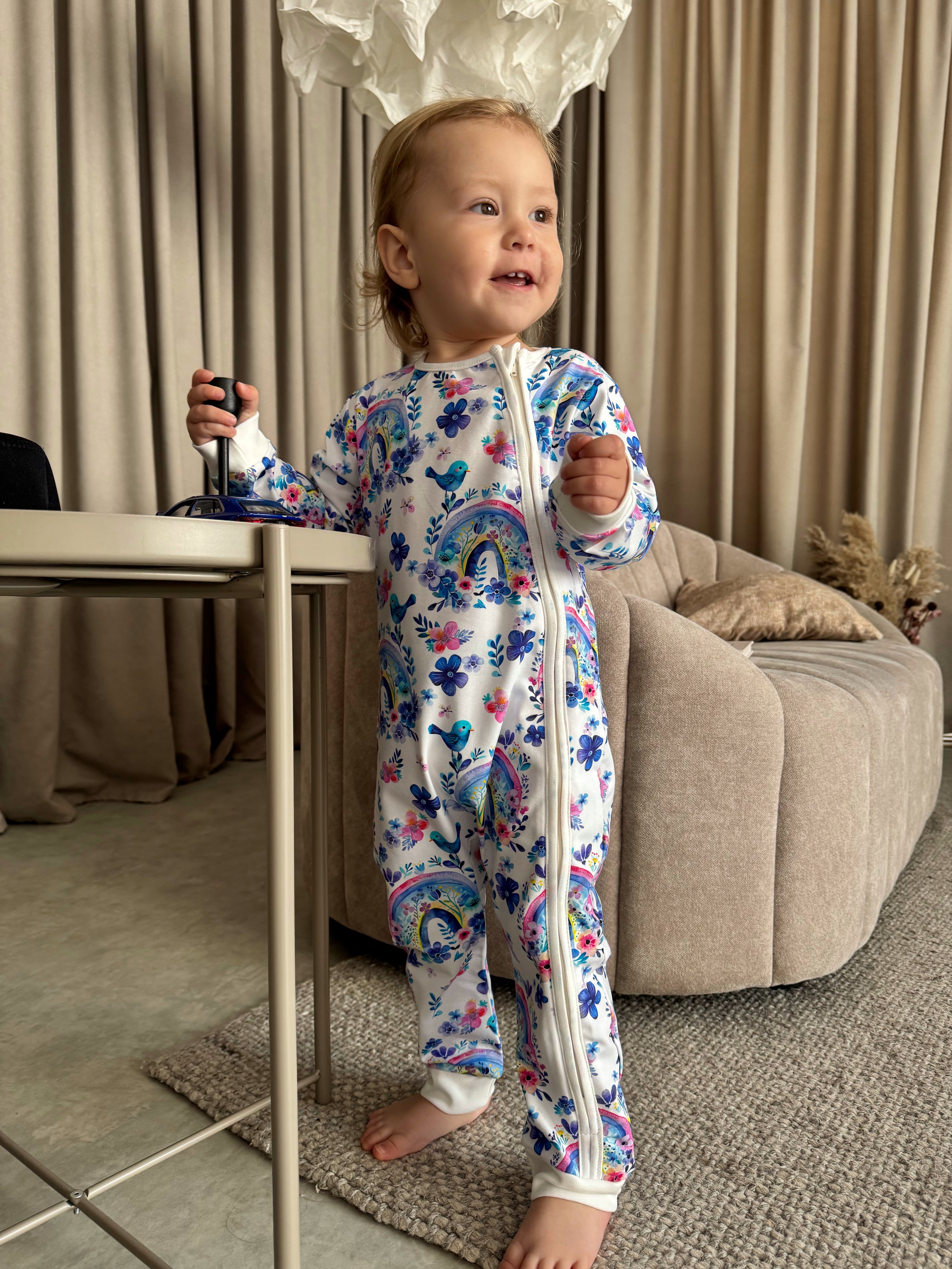 Child wearing a colorful onesie with a rainbow design in a living room.